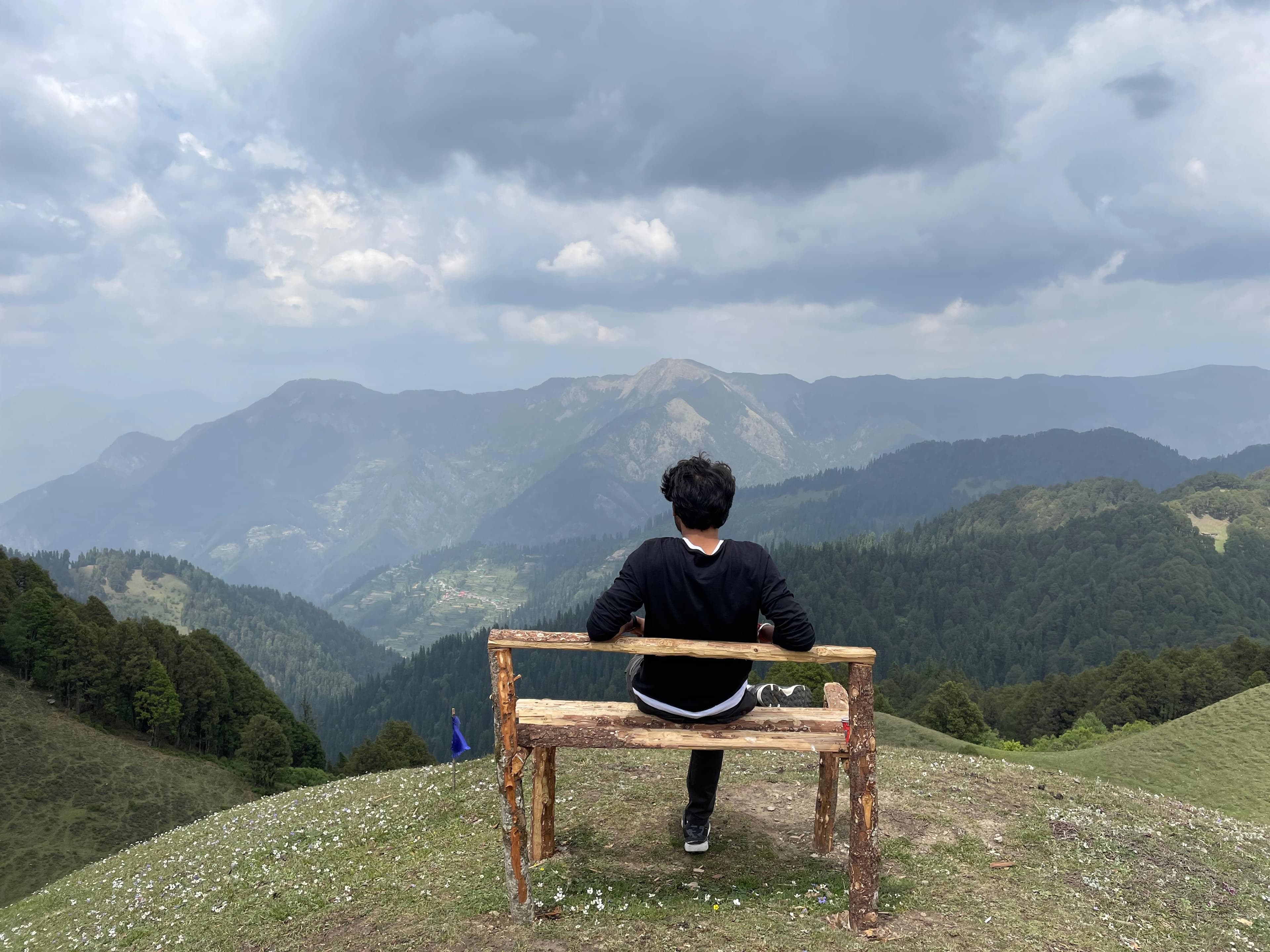 A traveller sitting on a wooden bench, looking out over forested mountain ridges near Jibhi, Himachal Pradesh